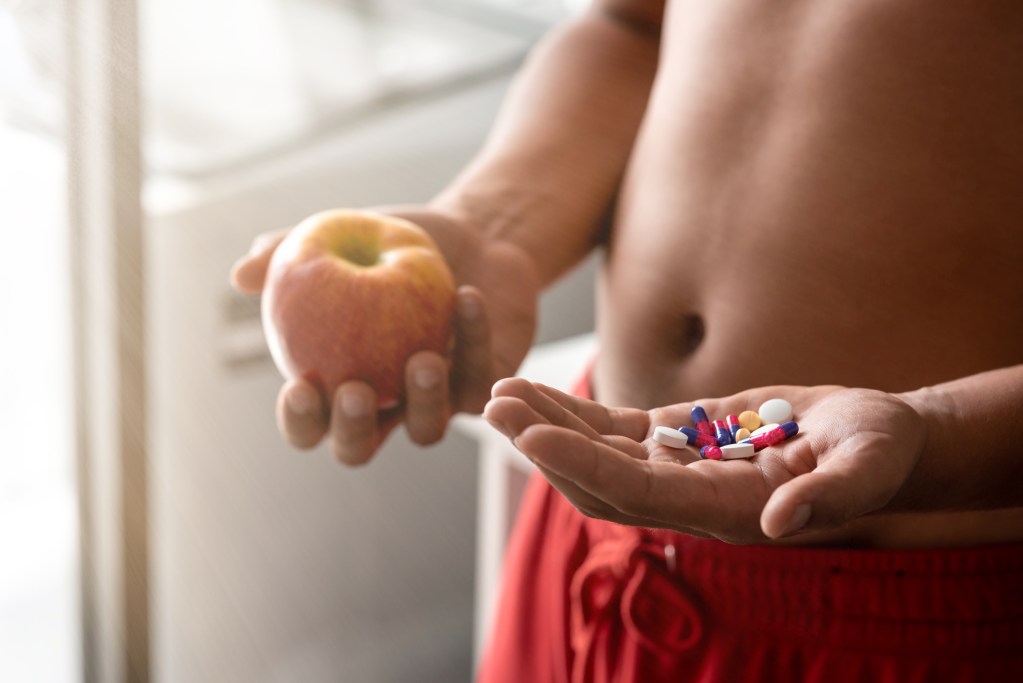 Close up of confused, puzzled man holding red fresh apple in one hand and pill, vitamins in another. He is trying to decide which choice is the best one. Horizontal shot